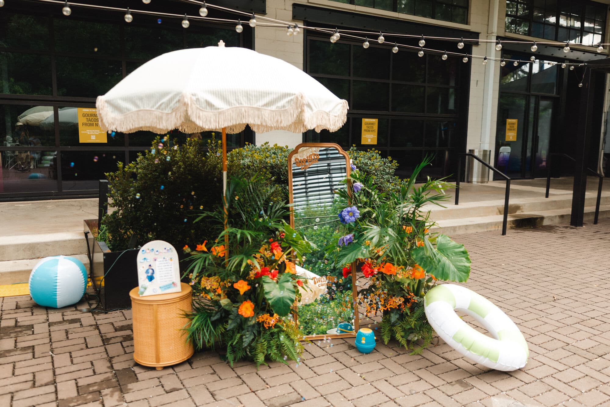 A beach-club photo moment styled by Twin Fins — fringed umbrella, tropical florals, and a mirror