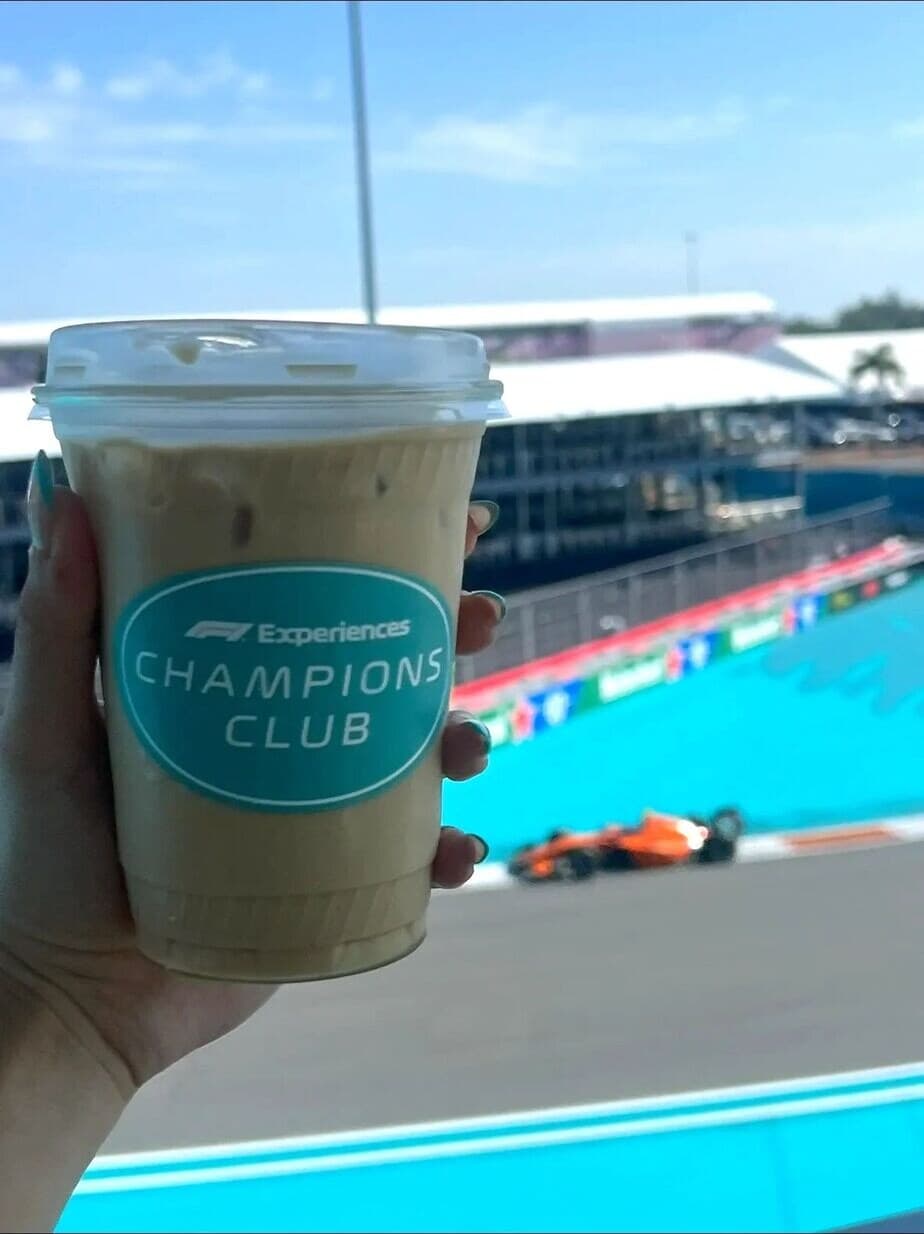 An iced latte in an F1 Experiences Champions Club cup, held up above the Miami circuit as a car passes