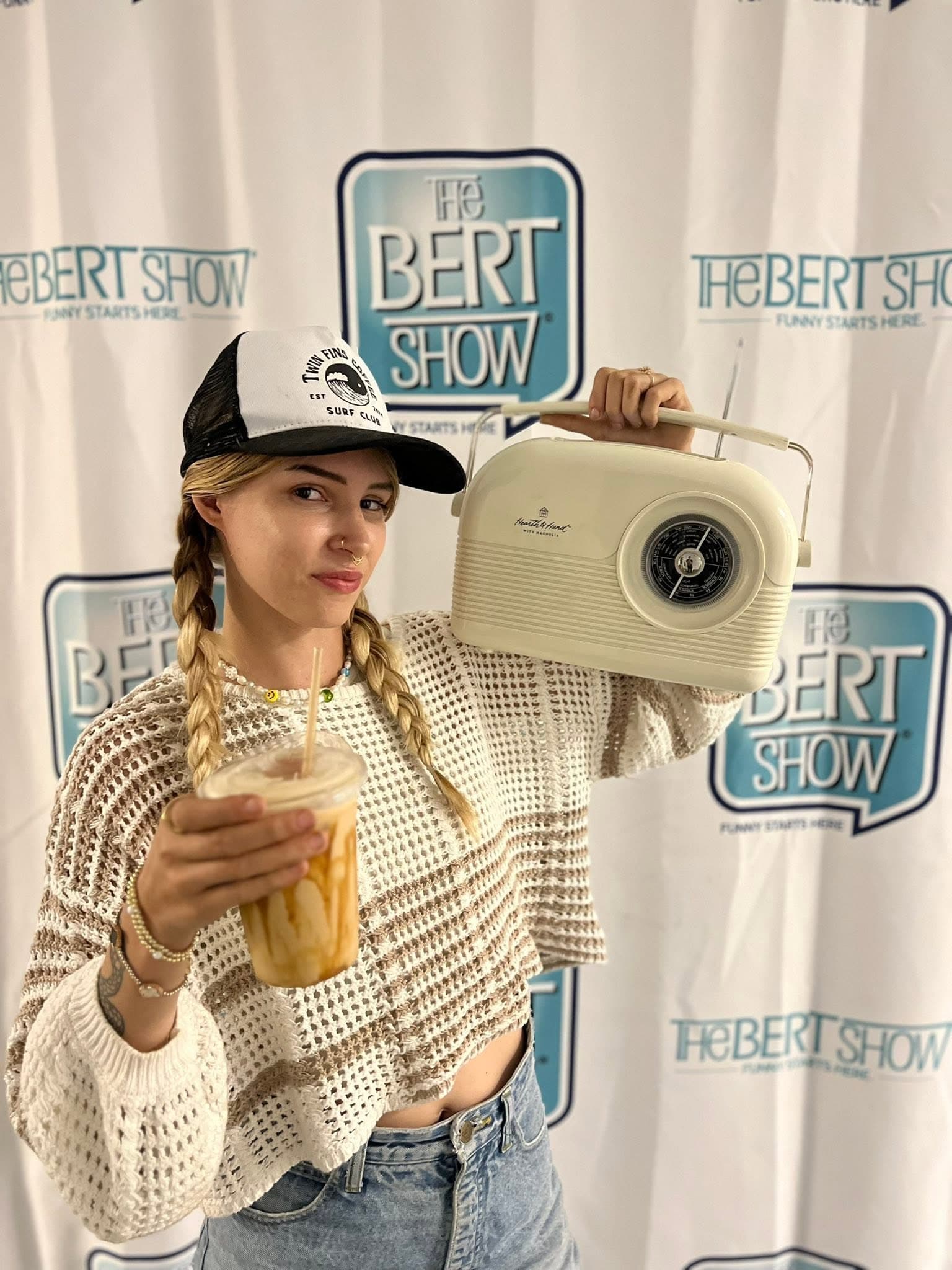 Natalia at The Bert Show step-and-repeat in a Twin Fins Surf Club hat, holding an iced coffee and a retro radio