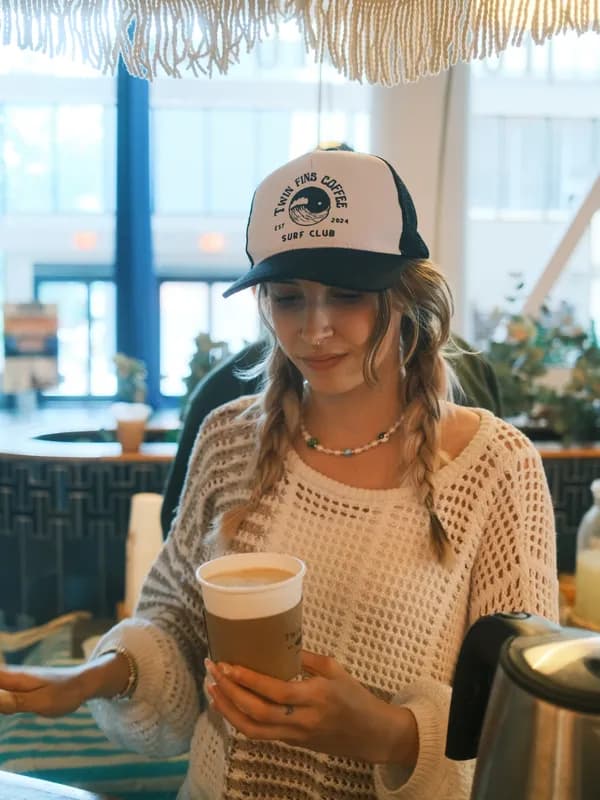 Natalia Tureta in a Twin Fins Surf Club trucker hat, holding a finished latte at the cart
